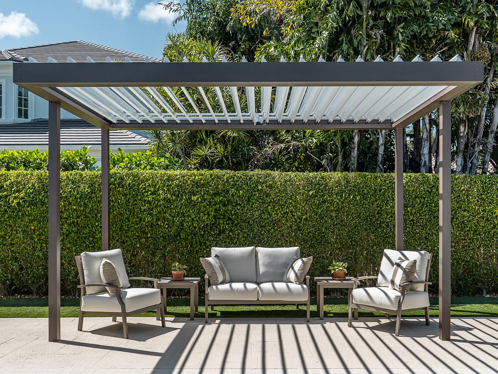 Freestanding louvered pergola with open blades casting striped shade over a lounge set in front of a tall hedge.