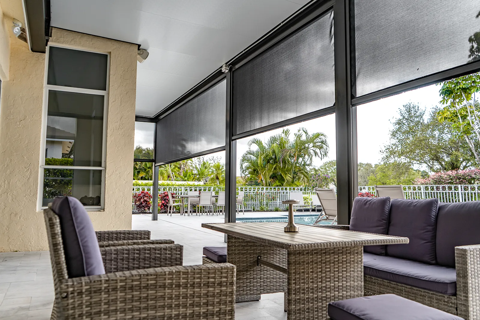 Motorized retractable screens half lowered on a covered patio, with wicker seating and palms beyond.