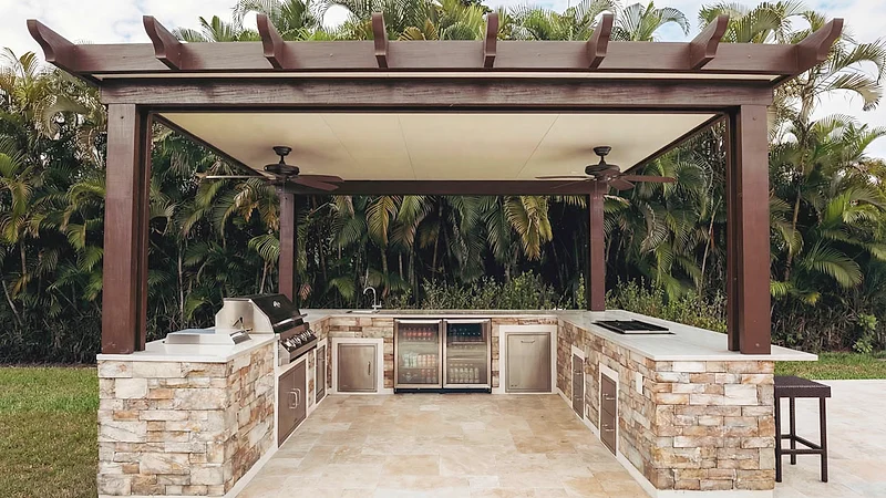 Brown pergola with ceiling fans over a U-shaped stacked-stone outdoor kitchen with a grill and drinks fridges, palms behind