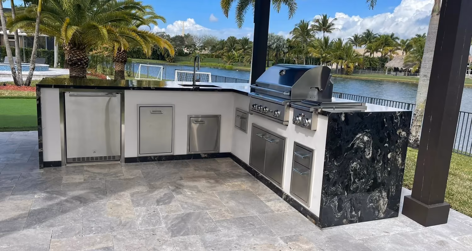 L-shaped outdoor kitchen with black granite waterfall under a dark pergola beside a lake.