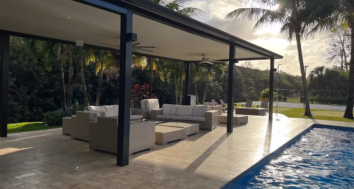 Black-framed insulated patio cover with ceiling fans and wicker sofas beside a lap pool at sunset
